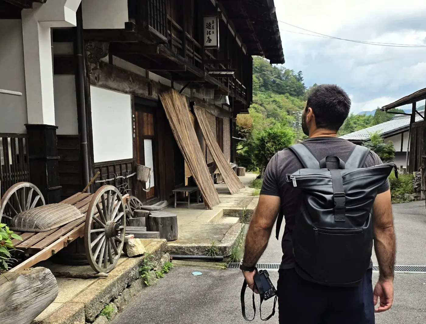 Gabriel, de espaldas con mochila y cámara, camina entre casas de madera tradicionales en la ruta Nakasendo